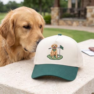 Op de afbeelding: Een crèmekleurige baseballpet met een donkergroene klep. Het voorpaneel is voorzien van een geborduurde afbeelding van een golden retriever met een groene bandana, die een golfclub in zijn bek houdt en op een green met vlag en golfbal zit.