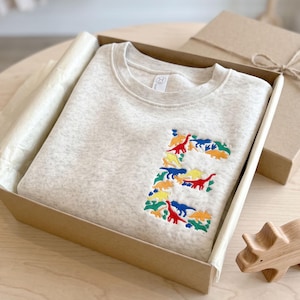May include: A light beige heathered crewneck sweatshirt featuring an embroidered letter E on the chest. The letter is composed of small, colorful dinosaur figures in shades of red, blue, yellow, orange, and green, neatly folded inside a cardboard box.