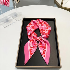 Peut inclure: Un foulard en soie rose et rouge à motifs de cœurs, présenté sur un plateau noir. Le foulard est noué au centre, avec les extrémités déployées. Le motif présente un motif de cœur répété.
