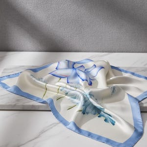 May include: A square, off-white silk scarf with a blue floral design and a light blue border. The scarf is draped on a white surface, showcasing the intricate floral pattern.