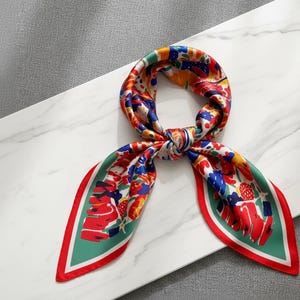 May include: A patterned silk scarf in a vibrant colour palette. The scarf displays a floral design with red, blue, yellow, and green tones. The edges are trimmed with a red border and green background. The scarf is tied in a knot.