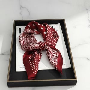 Peut inclure: Un foulard en soie bordeaux avec des pois blancs et des motifs cachemire. Le foulard est noué et présenté sur un plateau noir avec une bordure dorée, posé sur une surface blanche. Le motif du foulard est visible.