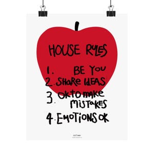 May include: A white poster featuring a large red apple graphic with black handwritten text that reads: HOUSE RYES, 1. BE YOU, 2. SHARE IDEAS, 3. OK TO MAKE MISTAKES, 4. EMOTIONS OK. The poster is suspended by two black metal clips at the top edge.
