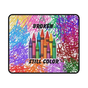 May include: A rectangular mousepad featuring a colorful design. The design includes a collection of crayons in various colors, with the words "BROKEN" and "STILL COLOR" in the center. The background is filled with scribbled lines in multiple colors.