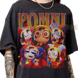 May include: A black short-sleeve t-shirt featuring a graphic print of five cartoon jester characters with red, blue, and yellow outfits. The text "POMNI" is printed in large, bold, orange-to-yellow gradient letters above the characters.
