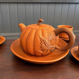 Può includere: Una teiera in ceramica a forma di zucca con finitura arancione testurizzata, dotata di un coperchio con stelo, un manico a forma di ramo e dettagli a foglia in rilievo. La teiera poggia su un piattino abbinato, circondata da tazze da tè.