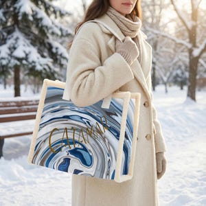 May include: A tote bag with a blue, white, and gray swirling pattern and the word "CATAMAR" in gold lettering. The bag has a cream-colored border and white straps. The background is a snowy winter scene.