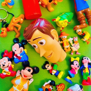 May include: A selection of brightly coloured cartoon character figurines, including Mickey and Minnie Mouse, Pluto, and Woody. The toys are arranged on a green surface, showcasing a variety of poses and outfits. The vibrant colours and familiar characters create a playful and nostalgic feel.