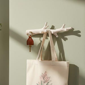 May include: A white, branch-shaped wall hook with a key and a tote bag hanging from it. The key has a brown leather fob. The tote bag is beige with a floral design. The wall is painted a light green.