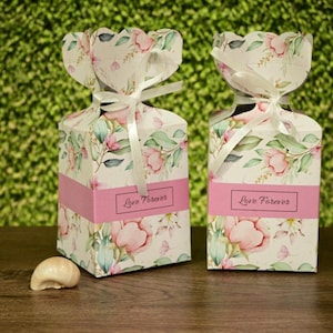 May include: Two floral gift boxes with pink accents and the text "Love Forever" on a pink band. The boxes are tied with white ribbons and sit on a wooden surface. The background is a green hedge.