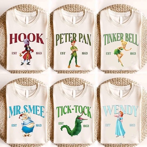 May include: Off-white t-shirts featuring characters from Peter Pan. Each shirt displays a character's name in a different color, along with an illustration and the text "EST 1953". Characters include Hook, Peter Pan, Tinker Bell, Mr. Smee, Tick-Tock, and Wendy.