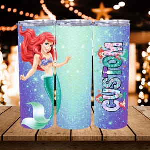 May include: Three stainless steel tumblers with a glittery design. One tumbler features Ariel from The Little Mermaid, another has a gradient, and the third says "CUSTOM" with mermaid graphics. The tumblers are on a wooden surface.