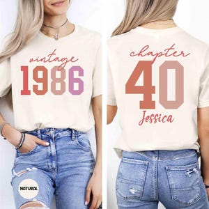 May include: Cream-colored t-shirt with the text "vintage 1986" on the front in shades of red and pink. The back of the shirt features "chapter 40 Jessica" in a similar color scheme. The shirt is paired with blue jeans.