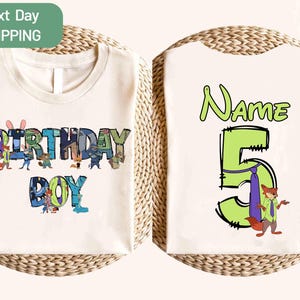 May include: Cream-colored t-shirt with a colorful "BIRTHDAY BOY" graphic on the front, featuring cartoon characters. The back of the shirt has a large green number "5" with a fox character and the word "Name" above it. The shirt is displayed on a woven surface.