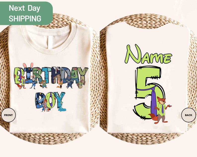 May include: Cream-colored t-shirt with a colorful "BIRTHDAY BOY" graphic on the front, featuring cartoon characters. The back of the shirt has a large green number "5" with a fox character and the word "Name" above it. The shirt is displayed on a woven surface.