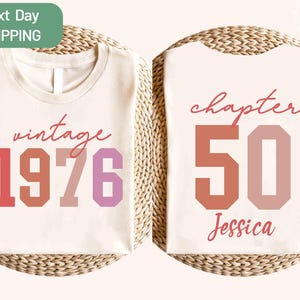 Puede incluir: Camiseta color crema con el texto "vintage 1976" en tonos rojos y rosas en la parte delantera. La parte trasera de la camiseta presenta "chapter 50" y el nombre "Jessica" en una combinación de colores similar. La camiseta se muestra sobre una superficie tejida.