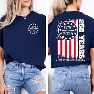 May include: Navy blue t-shirt with white graphic on the front and back. The front features the text "1776 2026" inside a circle of stars. The back displays a distressed American flag design with "250 YEARS" and "QUARTER-MILLENIAL" text.
