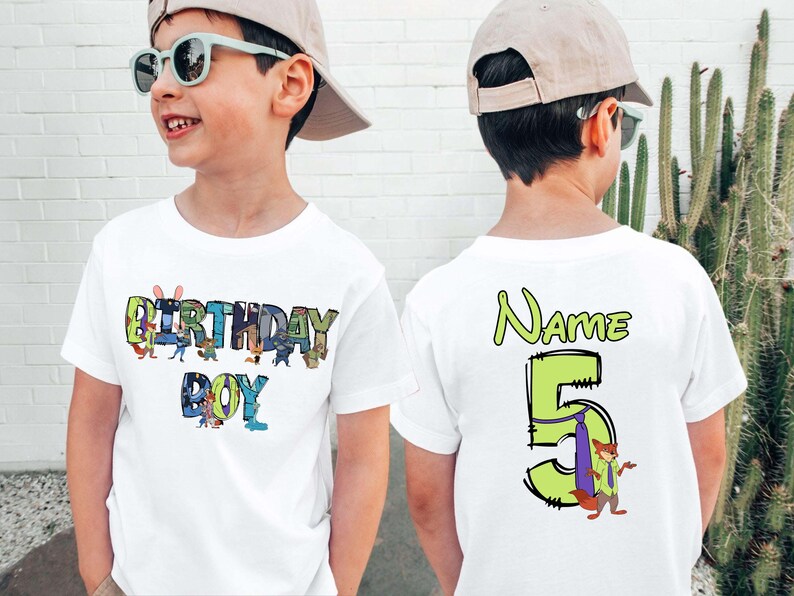 May include: White t-shirt with the words "BIRTHDAY BOY" in colorful letters and cartoon characters. The back of the shirt has the word "Name" and the number "5" with a cartoon fox. A fun birthday shirt.