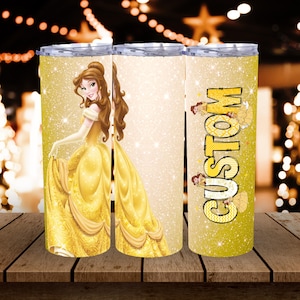 Peut inclure: Trois gobelets jaunes à paillettes avec un motif de princesse. L'un des gobelets représente une princesse en robe jaune, un autre est vierge et le troisième porte le mot "CUSTOM" en jaune. Les gobelets sont sur une surface en bois.