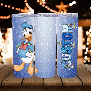 Op de afbeelding: Drie blauwe, met glitter bedekte roestvrijstalen bekers. Eén beker heeft Donald Duck, een andere is effen en de derde heeft het woord "CUSTOM" in blauw met Donald Duck-illustraties. De bekers staan op een houten oppervlak.