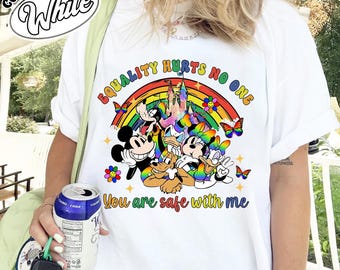 Op de afbeelding: Wit T-shirt met een kleurrijke afbeelding van Mickey Mouse, Minnie Mouse, Donald Duck en Pluto onder een regenboog. De tekst "EQUALITY HURTS NO ONE" en "You are safe with me" zijn ook afgebeeld.
