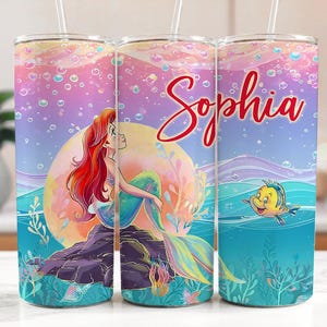 May include: A tumbler with a mermaid design and the name "Sophia" in red script. The tumbler has a colourful background with bubbles, a mermaid on a rock, and a yellow fish. The tumbler includes a straw.
