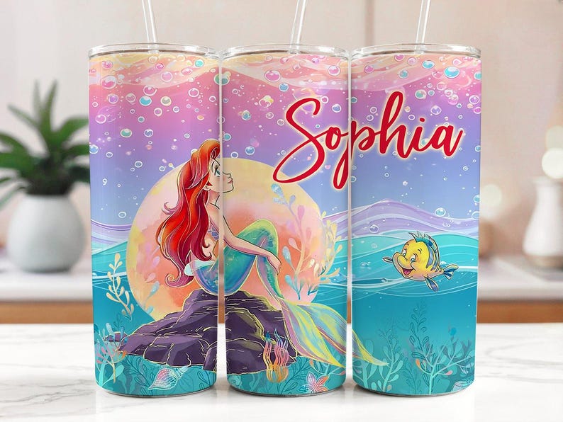 May include: A tumbler with a mermaid design and the name "Sophia" in red script. The tumbler has a colourful background with bubbles, a mermaid on a rock, and a yellow fish. The tumbler includes a straw.