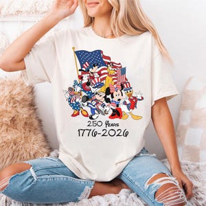 Puede incluir: Camiseta color crema con un dise&ntilde;o de personajes de Disney con atuendo patri&oacute;tico, sosteniendo banderas americanas. El dise&ntilde;o incluye el texto "250 Years 1776-2026". La camiseta es de estilo casual.