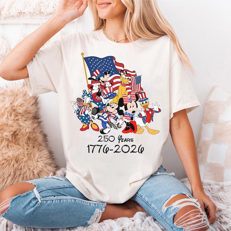Puede incluir: Camiseta color crema con un dise&ntilde;o de personajes de Disney con atuendo patri&oacute;tico, sosteniendo banderas americanas. El dise&ntilde;o incluye el texto "250 Years 1776-2026". La camiseta es de estilo casual.
