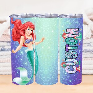 May include: A custom tumbler featuring a mermaid design. The tumbler has a glittery blue and purple background with a cartoon mermaid illustration. The word "CUSTOM" is vertically printed on the tumbler.
