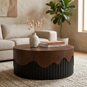 May include: A round coffee table with a dark wood top and a black, textured base. The table has a wavy design where the top meets the base. A vase with dried flowers and two books are on the table.