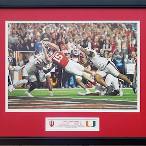 May include: Framed action shot of a football game, featuring players in red and white uniforms. The image is set against a red mat and black frame. A banner at the bottom reads "Unstoppable" with team names and scores.