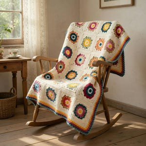May include: A cream-coloured crochet blanket with colourful floral squares draped over a wooden rocking chair. The blanket features red, yellow, blue, and orange flower designs. The chair is made of light-coloured wood.