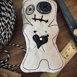 May include: A handmade, off-white fabric doll with black stitched details, including a heart, eyes, and mouth. A large black button serves as a head. Pins are inserted into the doll. A pair of black scissors and a ruler are in the background.