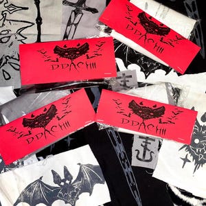 May include: Three rectangular red paper labels featuring a black graphic of a bat with wings spread and the text DDACHII, placed over various fabric patches with black and white screen-printed designs of swords, bats, and cross symbols.