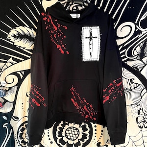 May include: A black hooded sweatshirt featuring a white rectangular patch on the chest with a black dagger illustration. The garment is decorated with red glittery splatter patterns across the front and sleeves, including a kangaroo pocket.