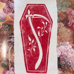 May include: A red coffin-shaped design with a white scythe and floral accents printed on a white fabric. The image has a vintage, gothic aesthetic. The fabric is likely a patch or a piece of material for crafting or apparel.