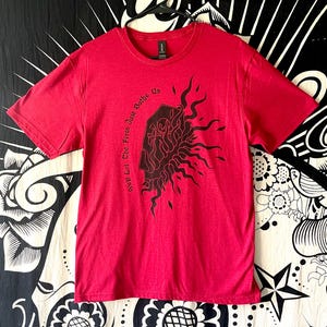 May include: A red t-shirt with a black graphic design featuring flames and the text "We'll Let The Fires Just Bathe Us". The shirt is hanging on a black hanger against a patterned background.