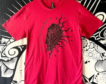 Puede incluir: Una camiseta roja con un diseño gráfico negro que presenta llamas y el texto "We'll Let The Fires Just Bathe Us". La camiseta está colgada en una percha negra sobre un fondo estampado.