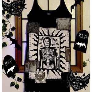 May include: A black tank dress featuring a central white rectangular patch with a graphic of a person with a skeleton torso surrounded by bats. The dress includes various smaller patches with graveyard and bat imagery, all attached with visible stitching.