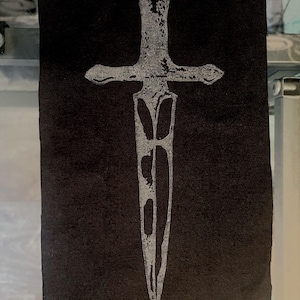 May include: Black rectangular patch featuring a silver dagger design. The dagger has a crossguard and a pointed blade. The design is centered on the patch, with the edges of the patch showing a raw, unfinished look.