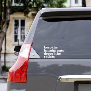 May include: A gray SUV rear window with a white decal. The decal reads "keep the immigrants deport the racists" in a sans-serif font. The vehicle's taillight is red and white.