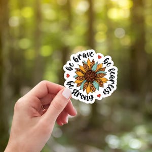 May include: A white sticker with a sunflower design and the words "be brave, be fearless, be strong". The sunflower has orange, yellow, and teal accents with a leopard print center and red heart details.