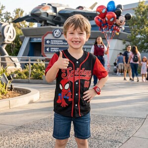 A young boy wearing a Spiderman jersey is smiling and giving a thumbs up, with a group of people and a Spiderman balloon in the background.