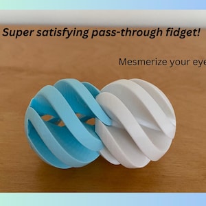 May include: Two spherical fidget toys, one light blue and one white, with a spiral design. The text "Super satisfying pass-through fidget!" and "Mesmerize your eyes" are visible. The toys are on a wooden surface.