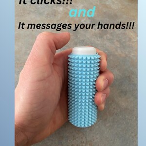 Puede incluir: Un masajeador de manos azul claro con una parte superior blanca y una superficie texturizada. El texto de la imagen dice "It clicks!!! and It massages your hands!!!"