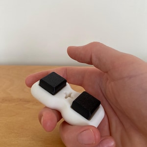 May include: A white ergonomic device with two black square pads. The device is held in a hand, showcasing its design and potential use. The pads appear to be textured, contrasting with the smooth white body.