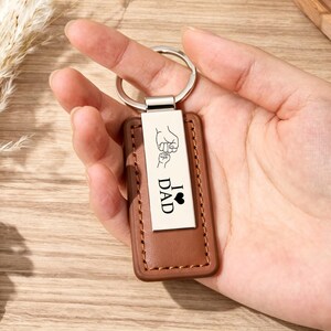 Puede incluir: Un llavero de cuero marrón con una placa de metal plateado. La placa tiene las palabras "I ♥ DAD" y un gráfico de la mano de un padre sosteniendo la mano de un niño. El llavero se sostiene en una mano.