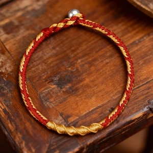 Puede incluir: Una pulsera trenzada con hilos rojos y dorados. La pulsera tiene una cuenta plateada en la parte superior y una sección dorada más gruesa en la parte inferior. Se muestra sobre una superficie de madera.