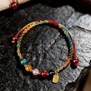May include: A colorful beaded bracelet with a gold charm. The bracelet features a braided design with red, yellow, green, and blue threads. It has several round, colorful beads and a small gold charm with Chinese characters.